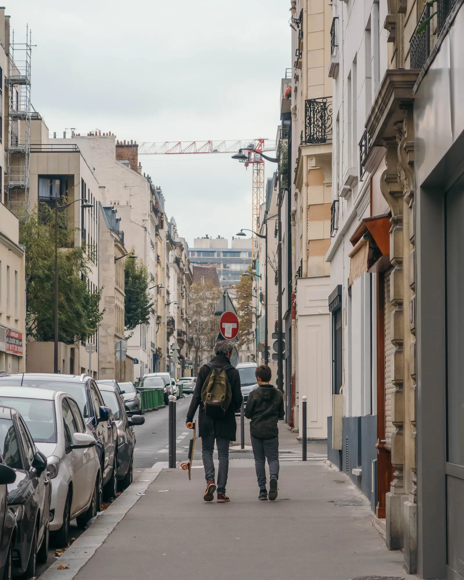 Two people walking along a narrow street of parked cars and pale stone apartment buildings.