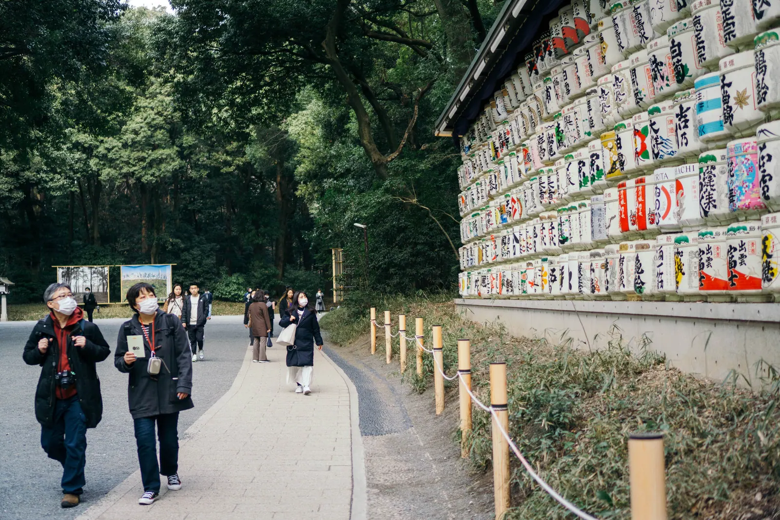 People walking a gravel path beside a wall of decorated sake barrels, forest behind.