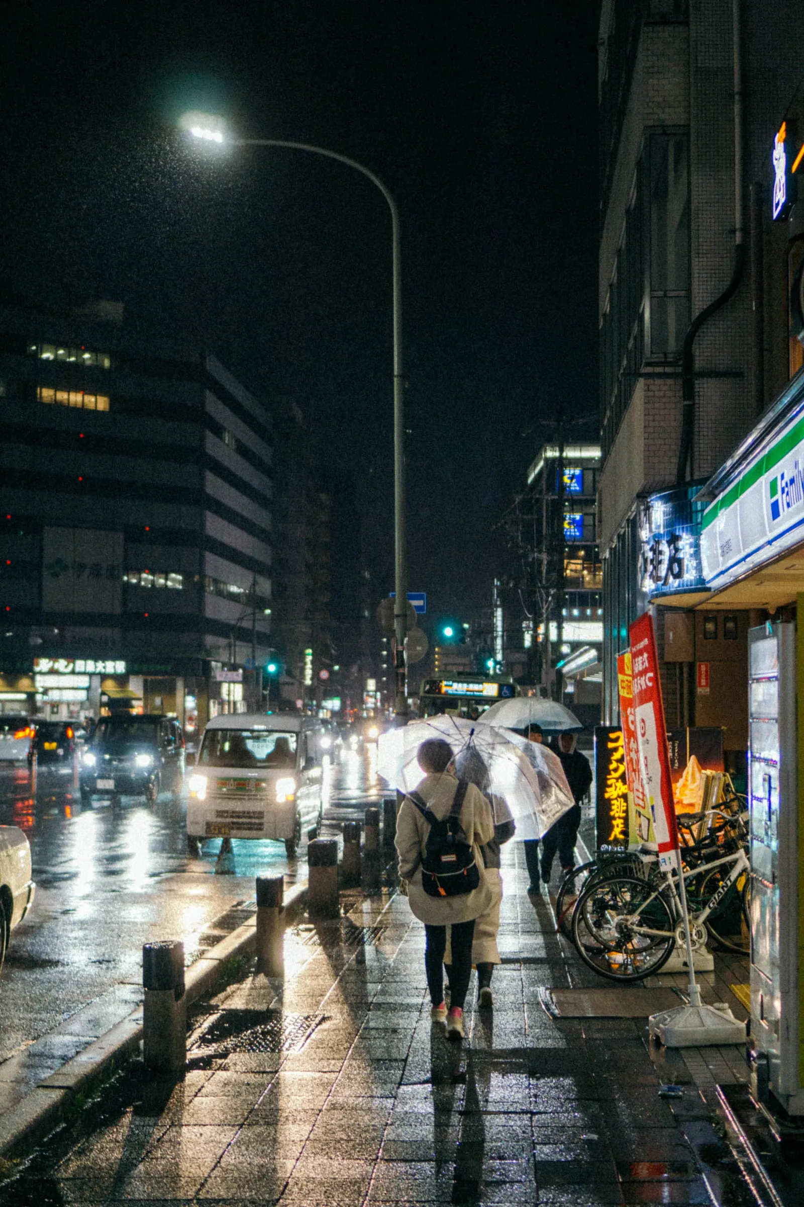 A rain-slick street at night, pedestrians with umbrellas passing a lit convenience store.