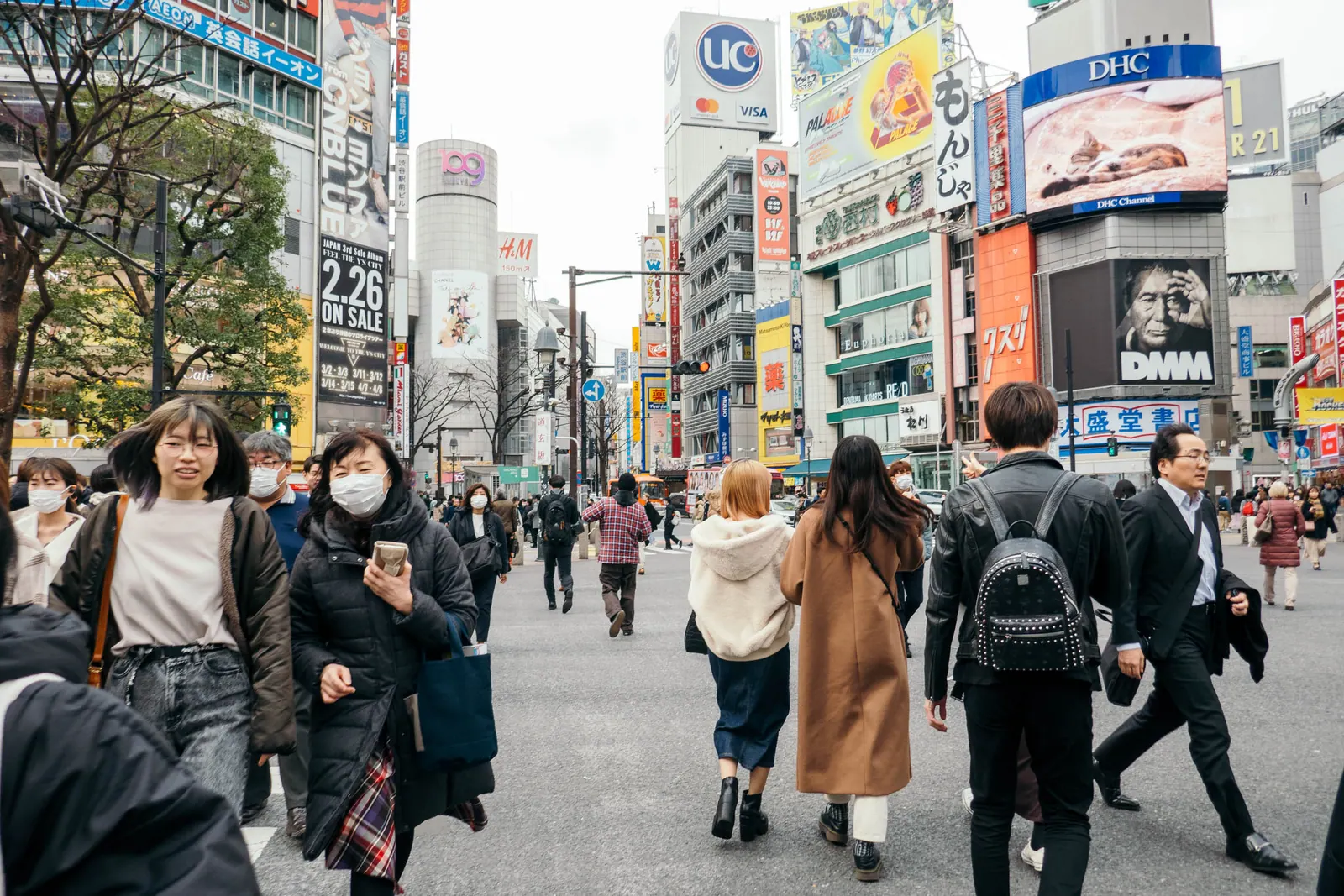 A crowd crossing a wide street between shops and billboards, several people wearing face masks.