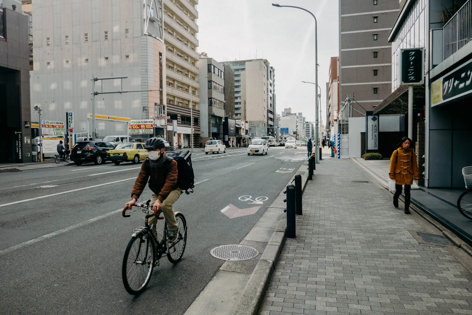 A masked cyclist riding down a wide, empty city street under an overcast sky.