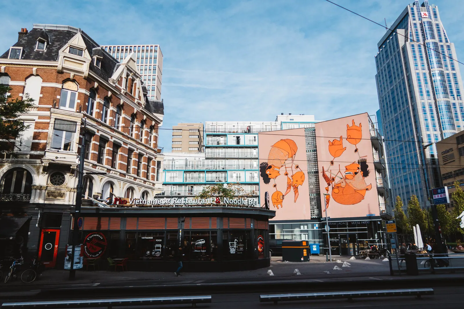A historic brick corner building beneath a glass tower and a large mural of orange figures.