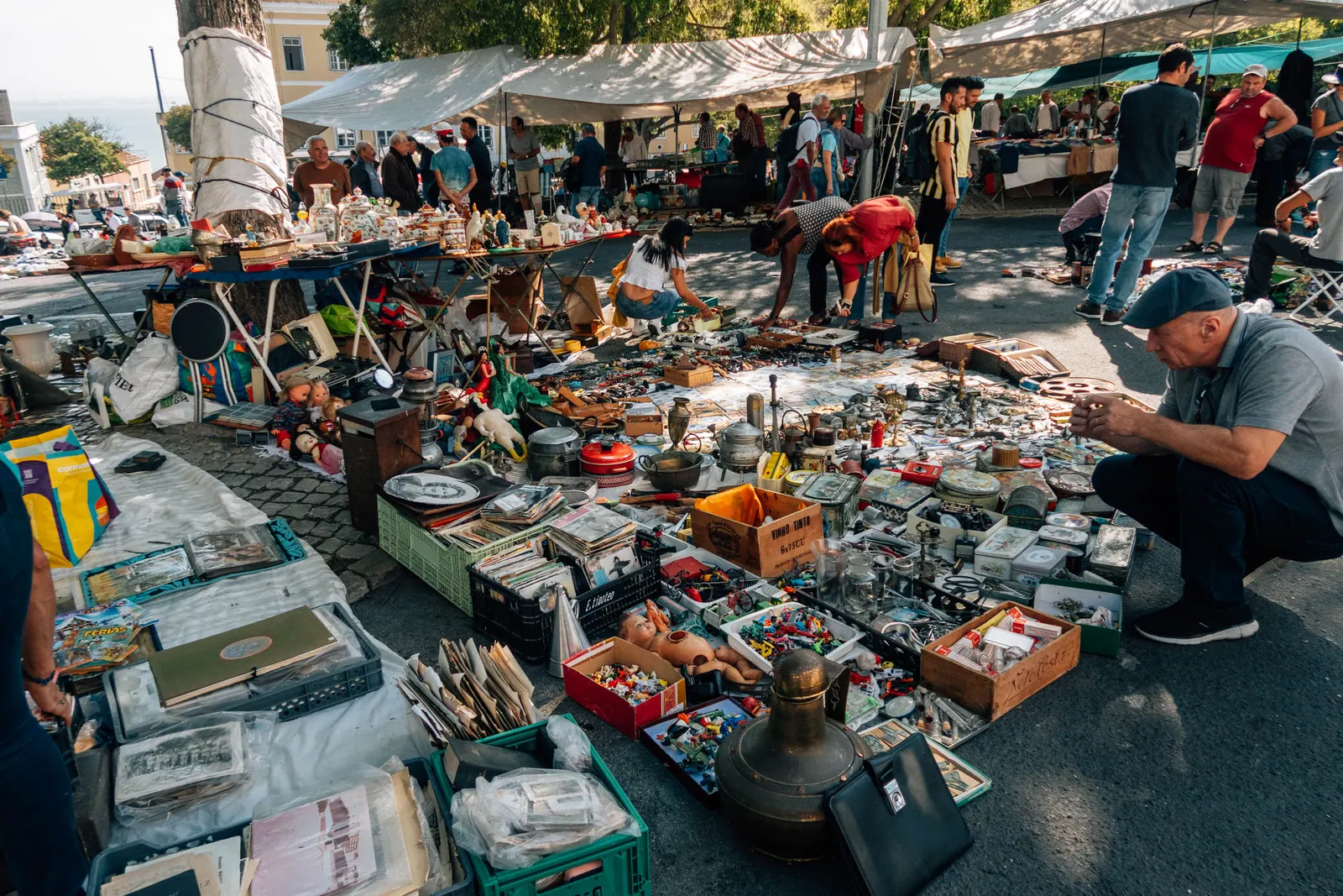 An open-air flea market, goods spread across blankets, a man crouching to look through them.