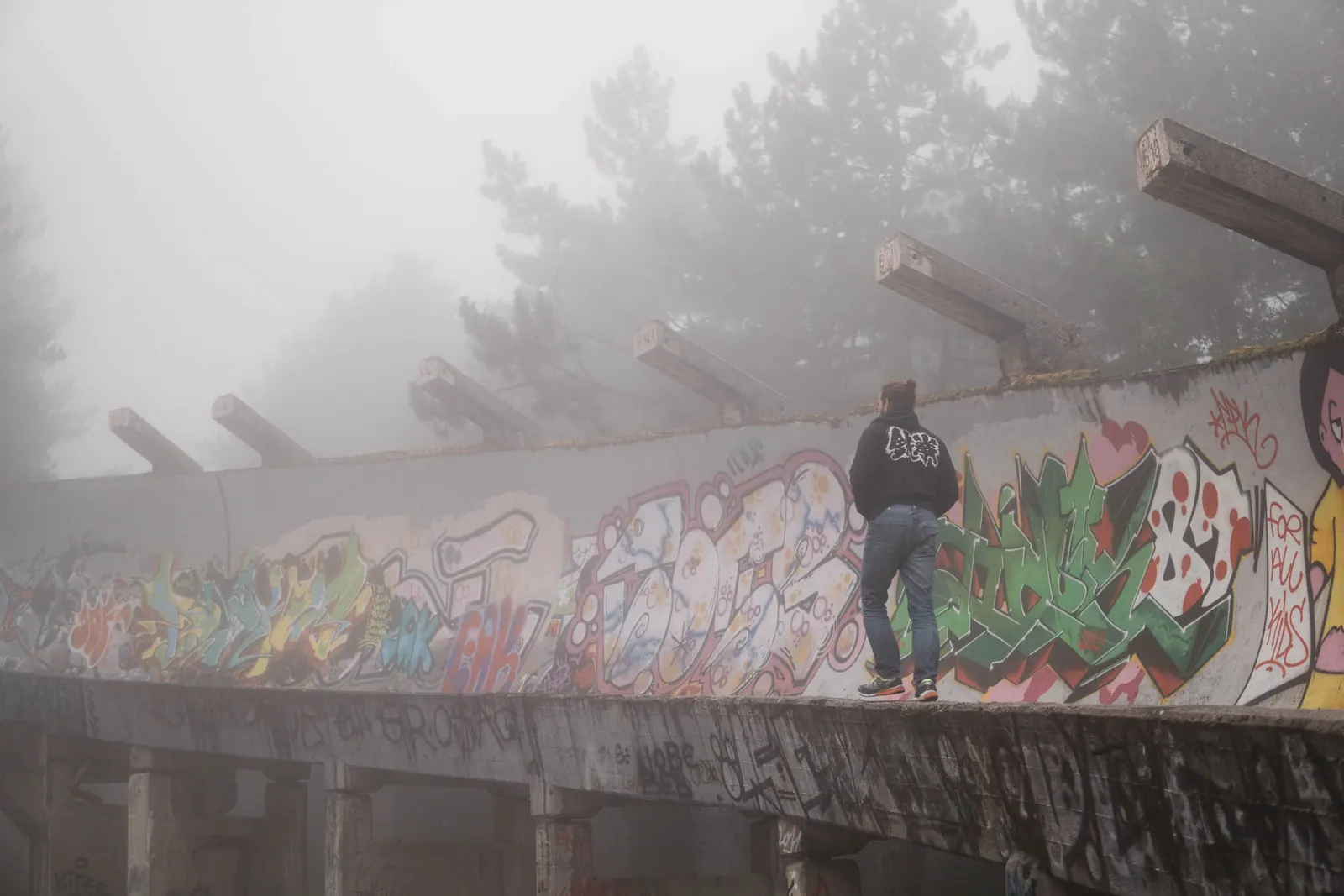 A figure in a hooded jacket standing on a graffiti-covered concrete track in dense fog.