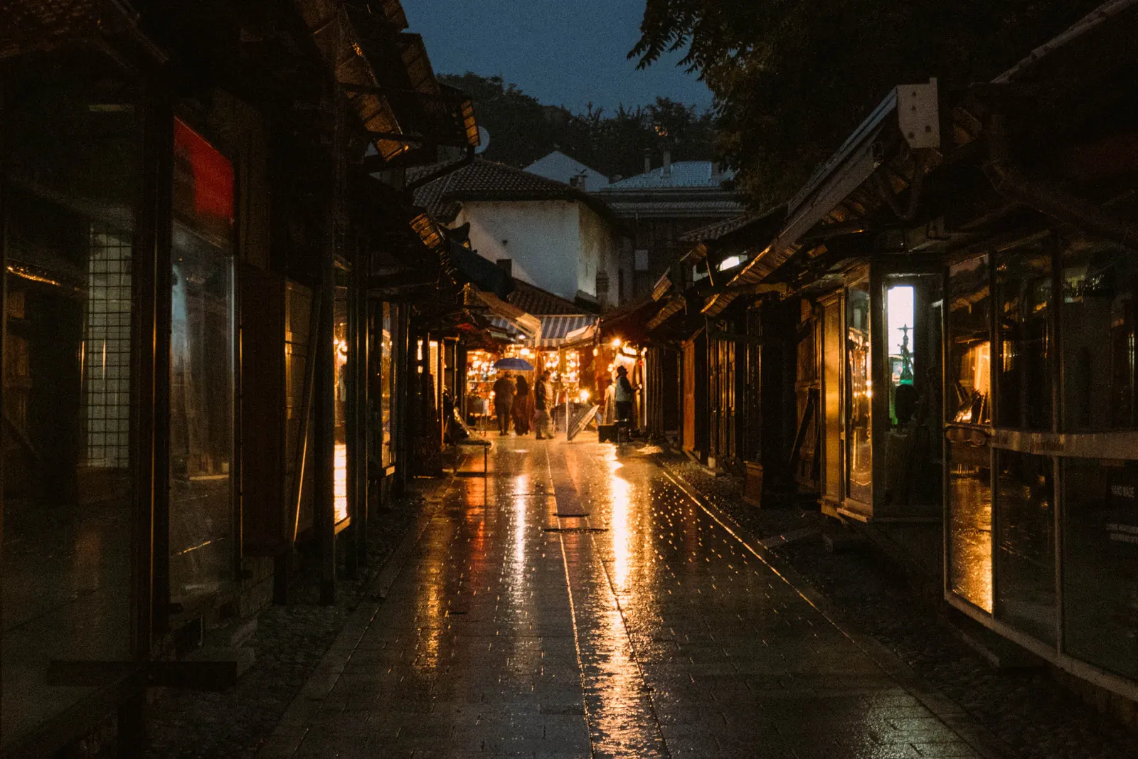 A narrow covered lane of wooden shopfronts at dusk, lamplight reflecting on wet paving stones.
